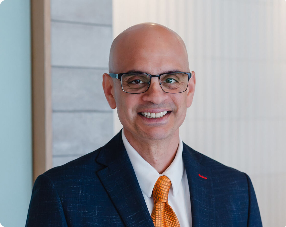 Apex Surgical doctor, Dr. Karim Al-Khatib, in a suit and orange tie smiling in a professional clinic setting.